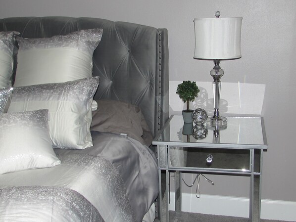Una cama bien hecha con un cabecero tapizado gris, una mesita de noche con una lámpara, jarrones decorativos y una pequeña planta.