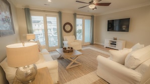 A spacious living room with a ceiling fan, a flat-screen TV, a white sofa, a coffee table, and a view of the outside through large windows. - BEST DEAL IN KURE BEACH!!! FULLY RENOVATED LUXURY HOME STEPS TO THE OCEAN & PIER (Kure Beach)