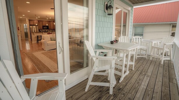 A covered porch with white wooden chairs and a table, a sliding glass door leading to a kitchen, and a teal-colored wall. - BEST DEAL IN KURE BEACH!!! FULLY RENOVATED LUXURY HOME STEPS TO THE OCEAN & PIER (Kure Beach)