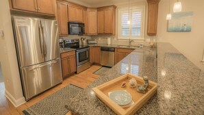A kitchen with wooden cabinets, stainless steel appliances, granite countertops, and a small island with a decorative bowl. - BEST DEAL IN KURE BEACH!!! FULLY RENOVATED LUXURY HOME STEPS TO THE OCEAN & PIER (Kure Beach)