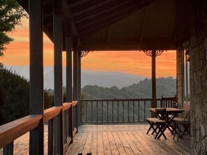 Een houten terras met een stenen muur en een leuning, met uitzicht op een bergachtig landschap tijdens de zonsondergang.