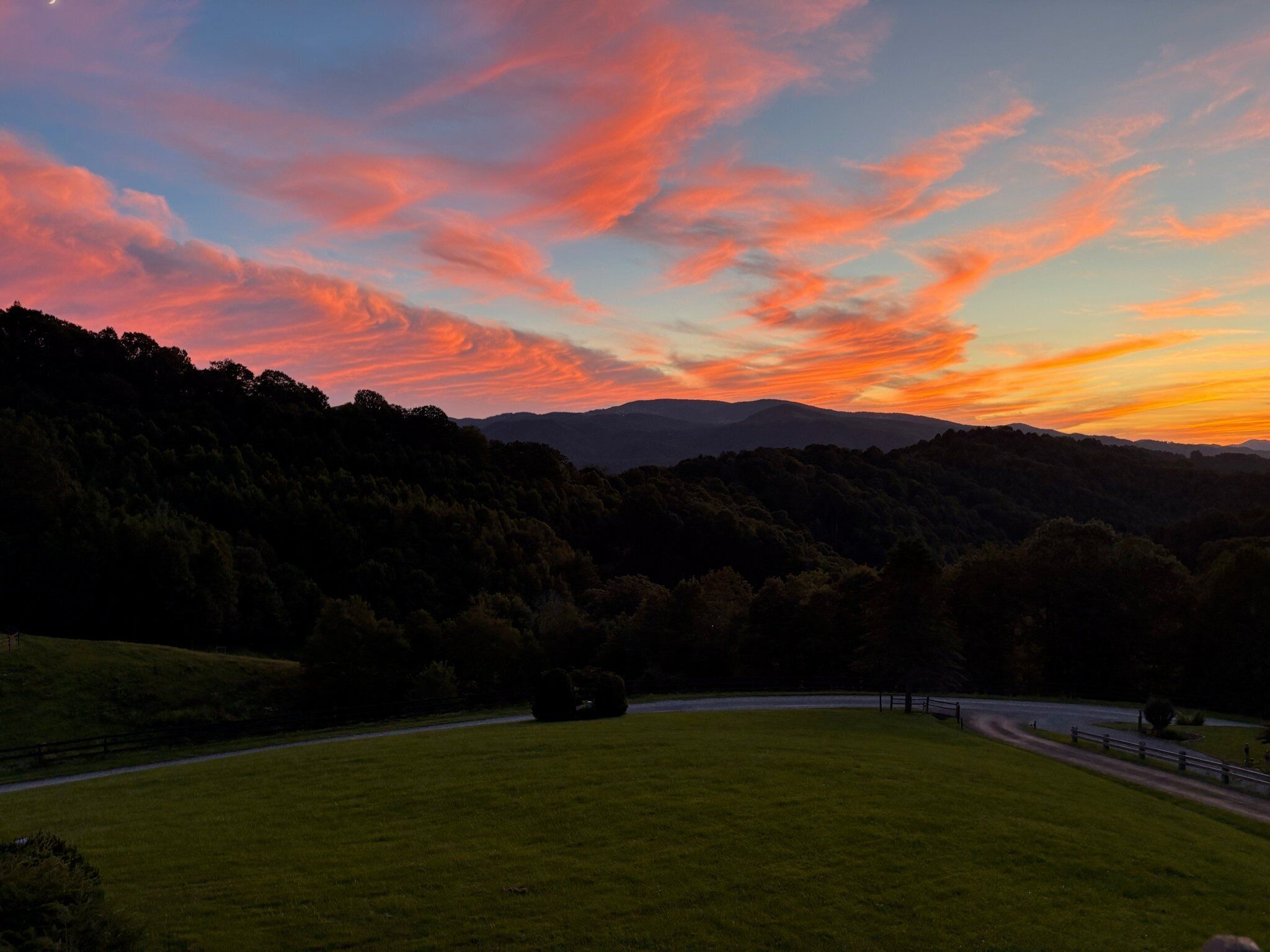 Ländliche Landschaft mit einer kurvenreichen Straße, grünen Feldern und einem bewaldeten Hügel im Hintergrund.