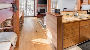 L’intérieur d’une cabane comprend un coin repas en bois, une cheminée, une cuisine avec un évier et une grande fenêtre donnant sur un paysage enneigé.