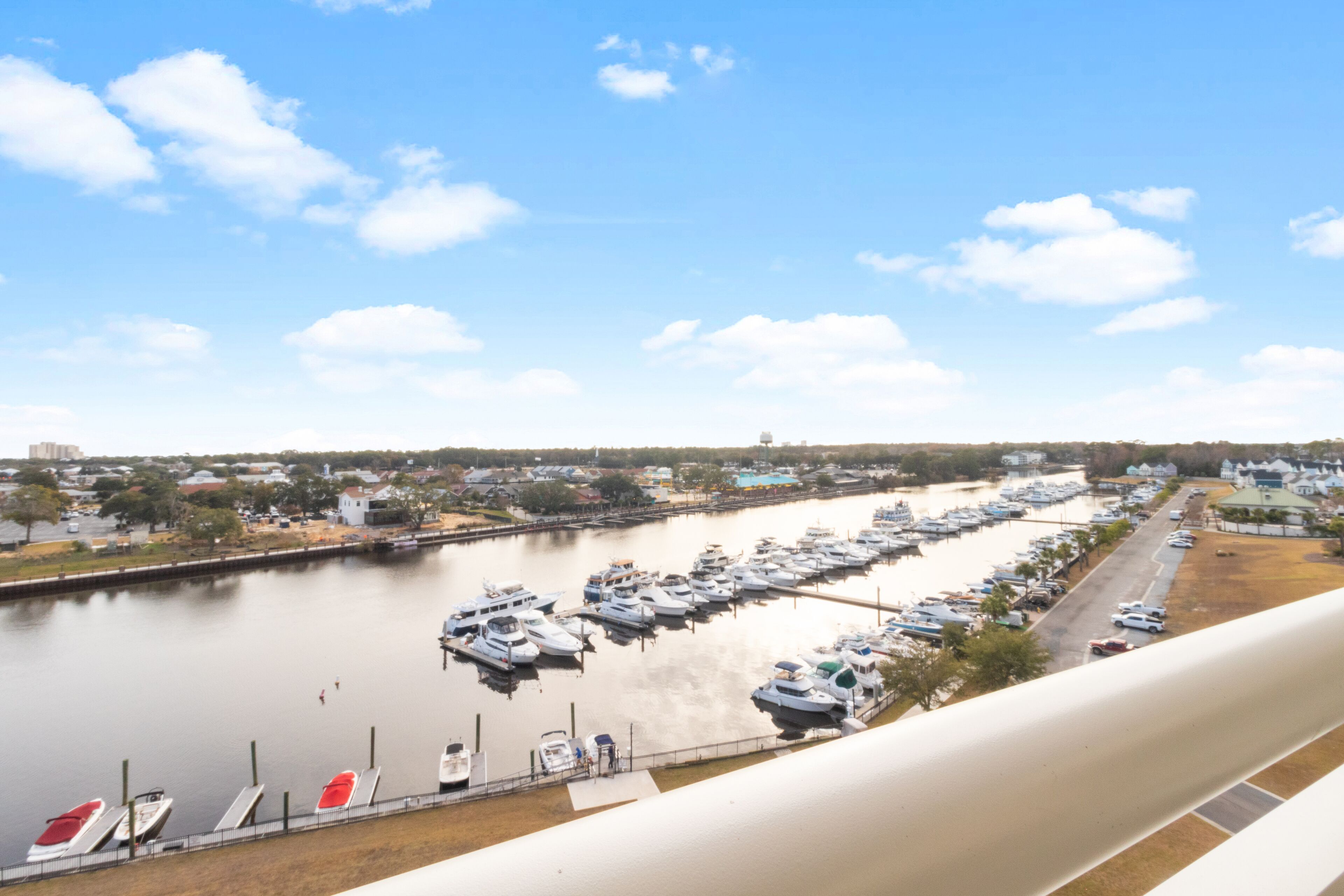 A marina with numerous boats docked along a waterfront, a road parallel to the water, and a clear sky.
