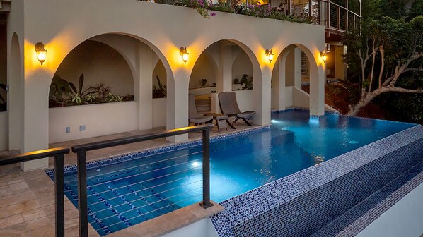 A swimming pool with a tiled edge, surrounded by a white wall with arched openings and wall-mounted lights.