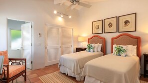 A hotel room with two beds, a desk, a chair, a ceiling fan, and three framed pictures on the wall.