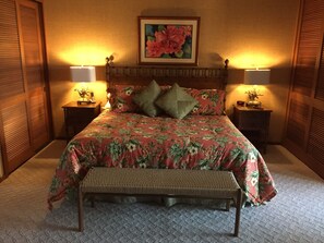 A bedroom with a floral bedspread, two bedside lamps, a wooden headboard, a bench, and a framed floral painting on the wall.