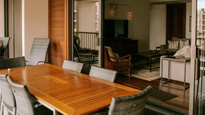 Uma área de refeições com mesa de madeira e cadeiras de vime, um ventilador de teto e vista para a sala de estar através de portas de vidro.