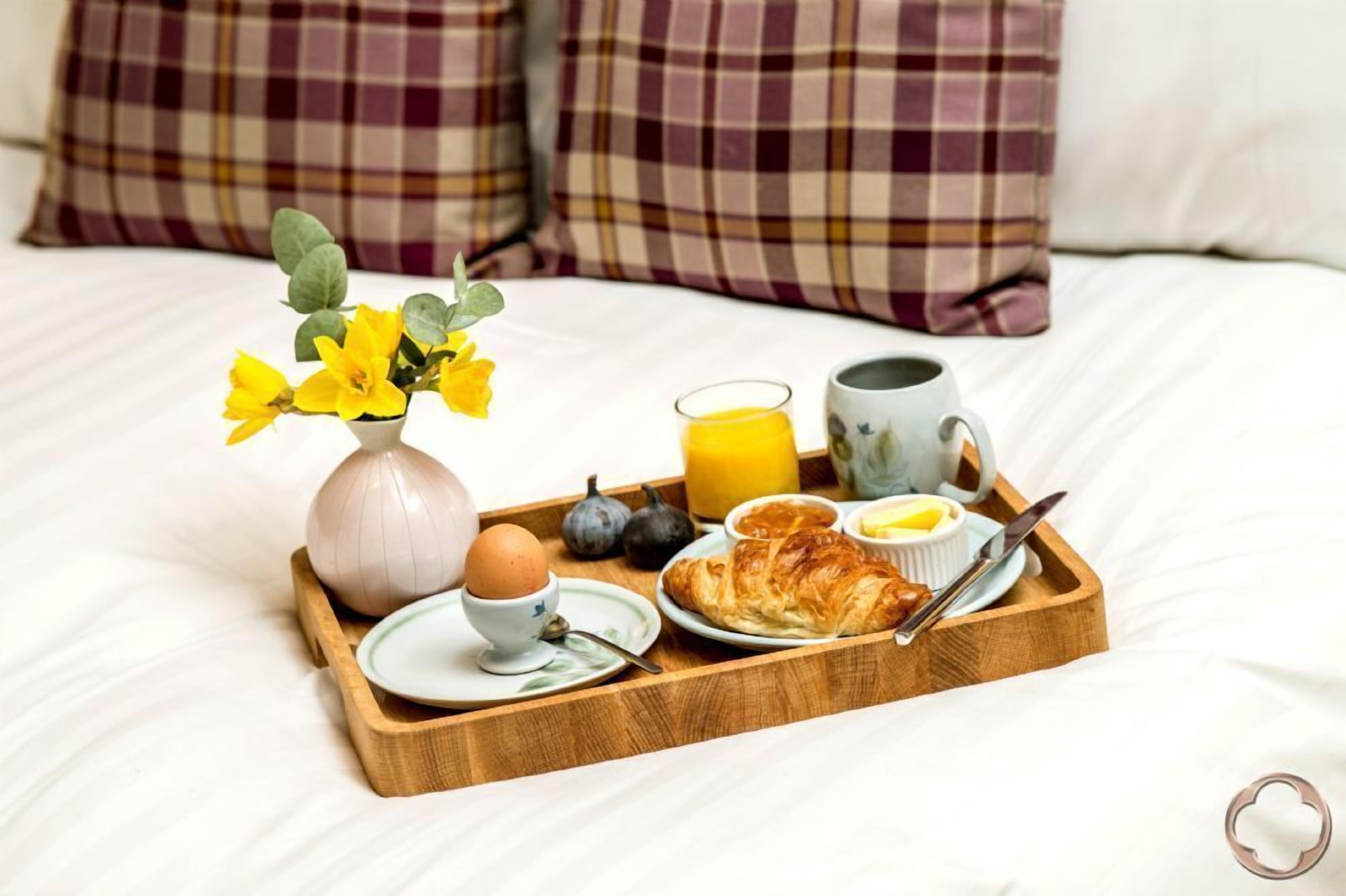 A breakfast tray on a bed with a vase of yellow flowers, a glass of orange juice, a mug, a croissant, and a plate with a boiled egg and a small bowl of butter.