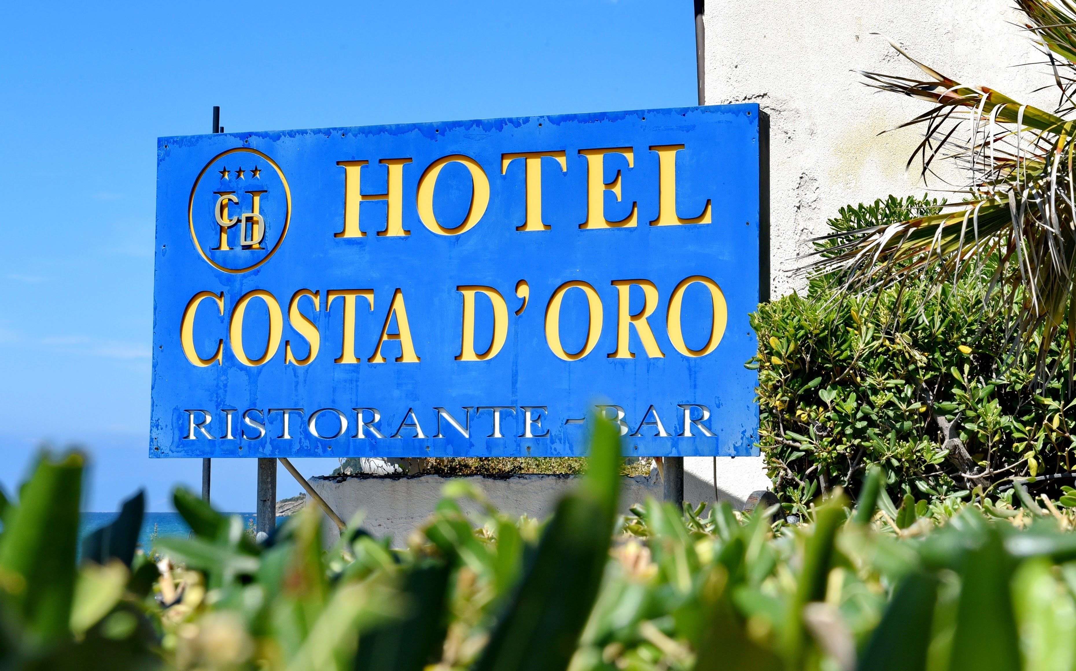 Ein blaues Schild mit goldener Schrift für das Hotel Costa d'Oro, das ein Restaurant/eine Bar anzeigt.