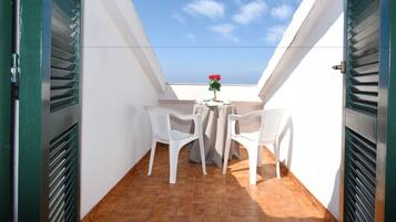 Un piccolo balcone con due sedie bianche di plastica, un tavolo, una pianta in vaso e un cielo azzurro sullo sfondo.