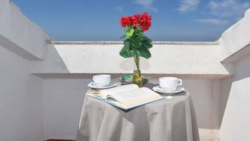 Ein kleiner Tisch mit einem Buch, zwei Tassen und einer Vase mit roten Blumen auf einem Balkon mit weißen Wänden und blauem Himmel.