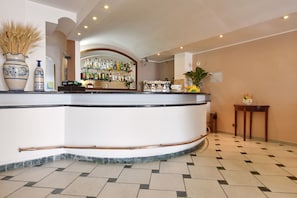 A hotel lobby with a curved reception desk, a bar area with various bottles, a large vase with dried plants, and a small table with a clock and flowers. - Costa d'Oro (Castellabate)