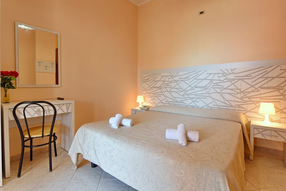 A hotel room with a bed, two bedside lamps, a desk with a chair, and a large abstract wall art piece.