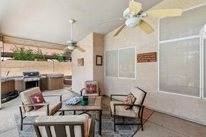 An outdoor patio with a grill, seating area, ceiling fans, and a large window with a view. - Lovely home in a quiet neighborhood with private pool! (Peoria)