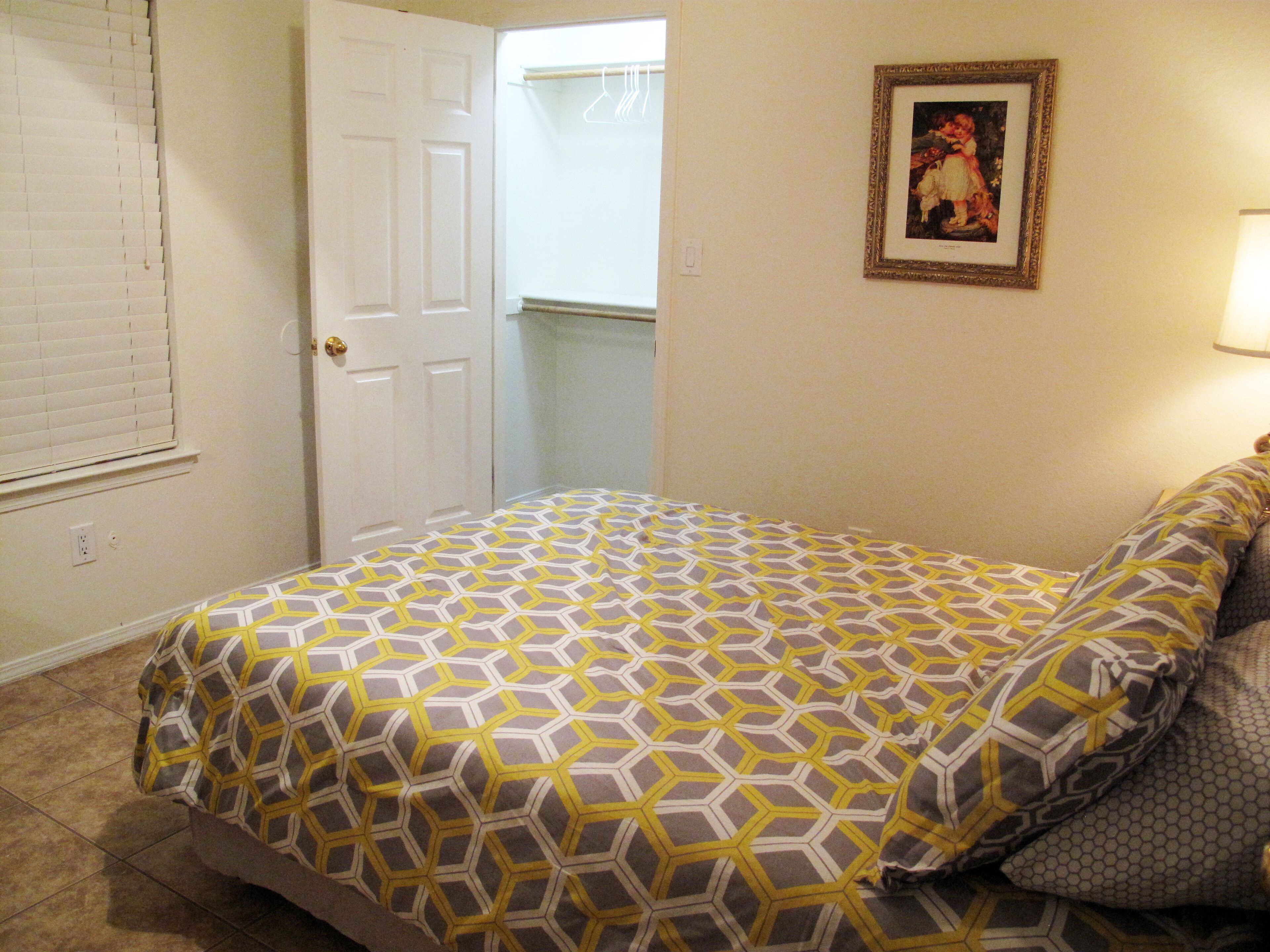 A bedroom with a patterned bedspread, a door, a framed picture on the wall, and a lamp.