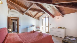 A bedroom with a wooden ceiling, a bed with a pink quilt, a white dresser, and a small table with a lamp.