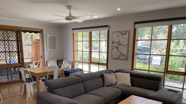 A modern living room with a grey sectional sofa, a wooden coffee table, and a dining area with a table and chairs. Large windows provide natural light and views of the outside. - Cowes Chapel Beach House (Cowes)
