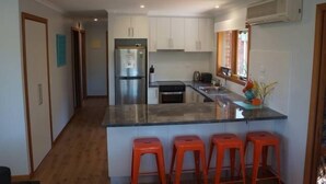 A modern kitchen with white cabinets, a stainless steel refrigerator, a built-in oven, and a central island with four red stools. - Cowes Chapel Beach House (Cowes)