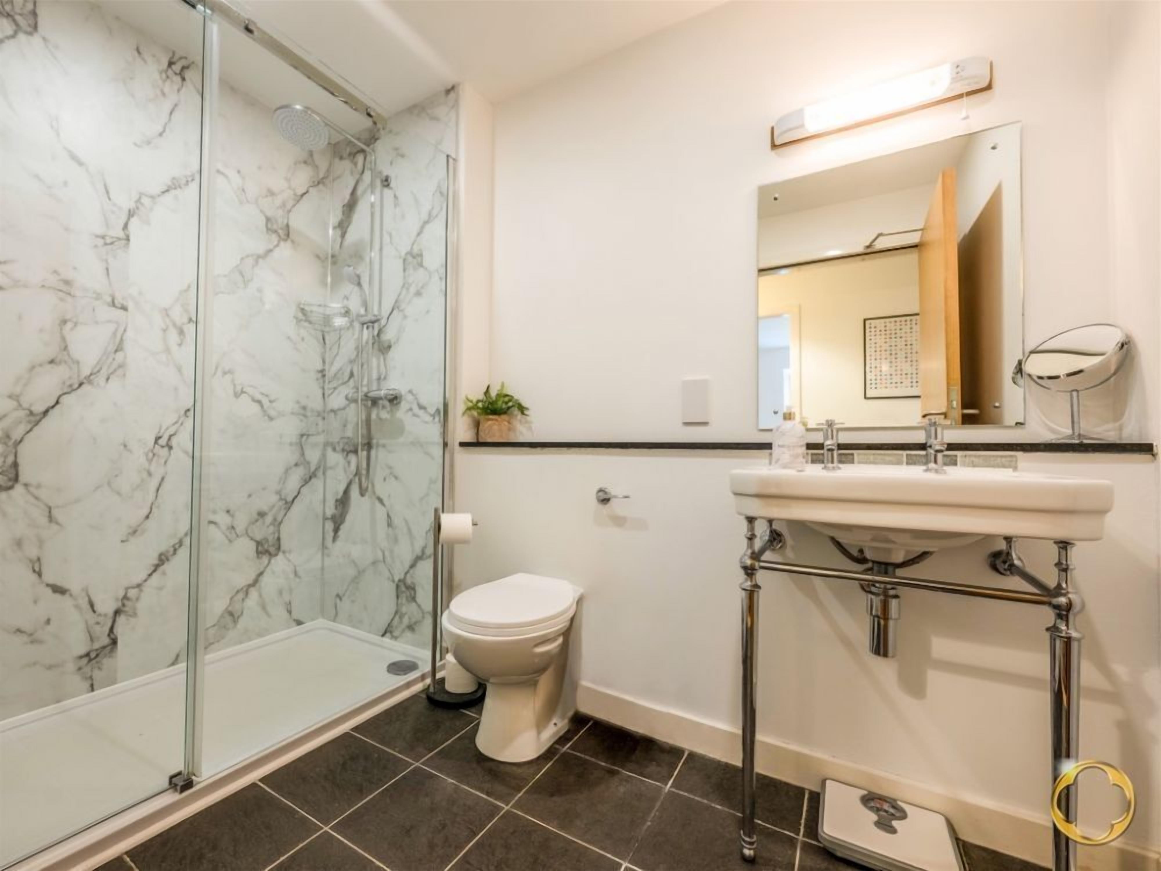 Ein Badezimmer mit einer Glaskabine zur Dusche, einer weißen Toilette, einem Waschbecken mit Spiegel und einer Marmorwand.