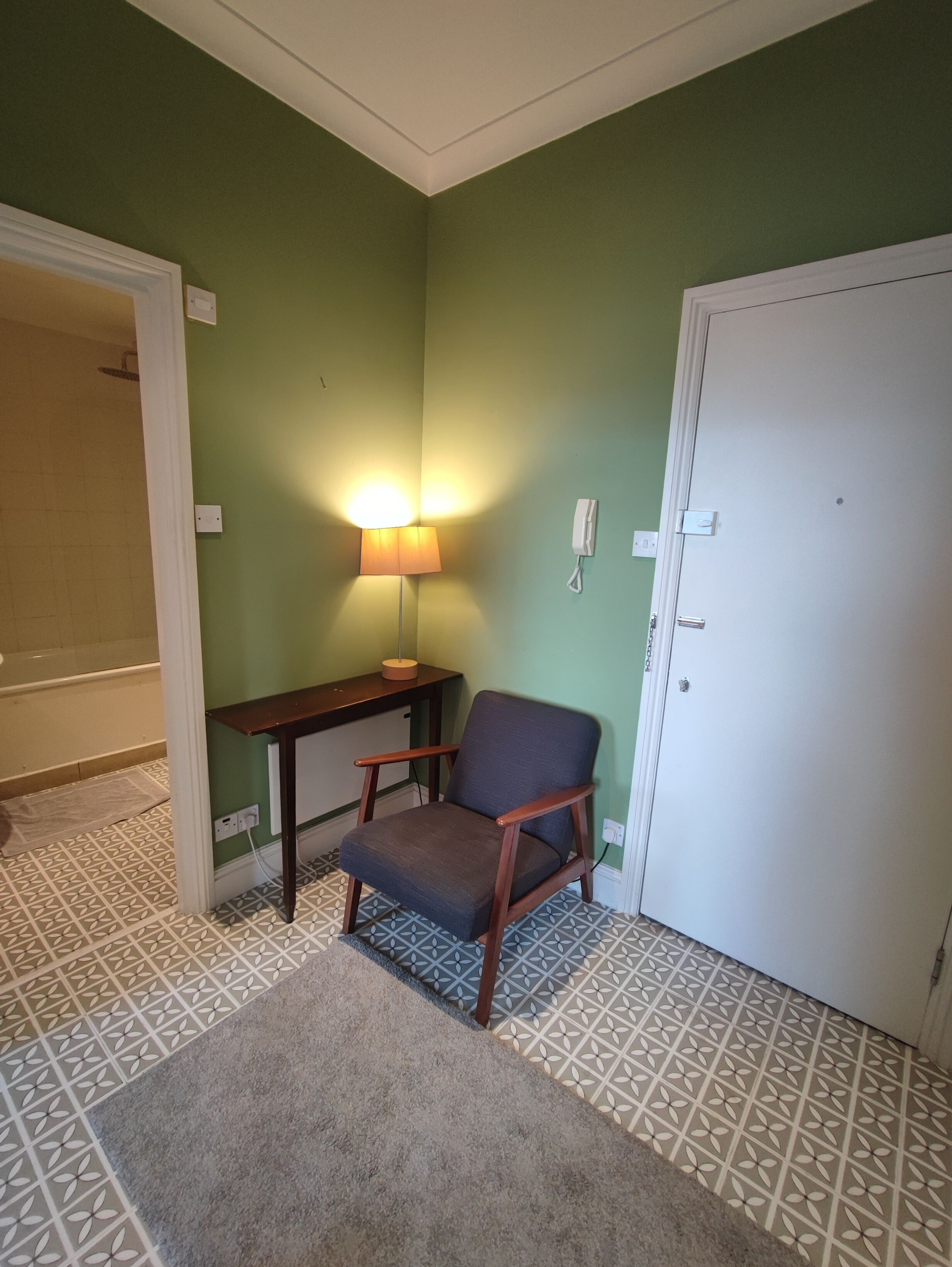 Una habitación con pared verde, una silla de madera, una mesita con lámpara, suelo estampado y una puerta blanca.