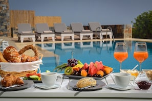 Colazione con croissant, frutta e bicchieri di succo, con poltrone a bordo piscina sullo sfondo.