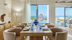 A dining area with a wooden table set for six, surrounded by cushioned chairs. A large window offers a view of the sea and a hammock. - Villa Greece Privat Seaview Villa with private heatable Pool  (Hersonissos)