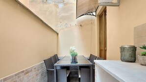 Un tavolo con piano in vetro, sedie in vimini, una pianta in vaso e una urna decorativa su un balcone con parete in mattoni e porta in legno.