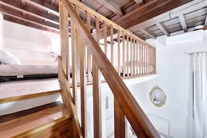 Un escalier en bois mène à une chambre située en mezzanine, qui comprend un matelas et un mur blanc.
