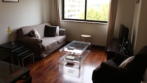 Una sala de estar con un sofá, una mesa de centro de vidrio, un televisor y una ventana con vistas a edificios.
