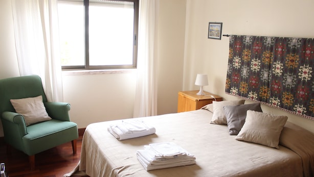 Una cama bien hecha con toallas dobladas, un sillón verde, una mesita de noche con lámpara y una ventana con cortinas.