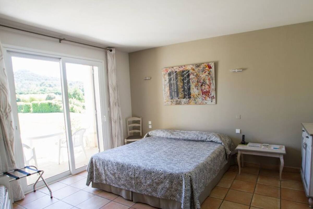 A bedroom with a bed, a small bedside table, a chair, a desk, and a view of the outdoors through a sliding door.