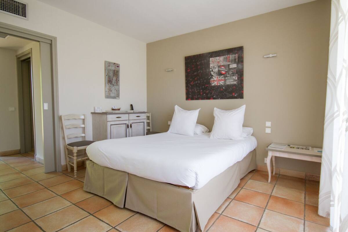 A hotel room with a large bed, a desk with a chair, a small table, and a wall-mounted artwork.
