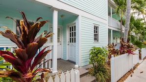 Una casa de dos pisos con un porche, cerca viva blanca y plantas tropicales.