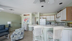 Una cocina moderna con armarios blancos, una isla central con taburetes altos y un comedor con sofá azul y sillón estampado.