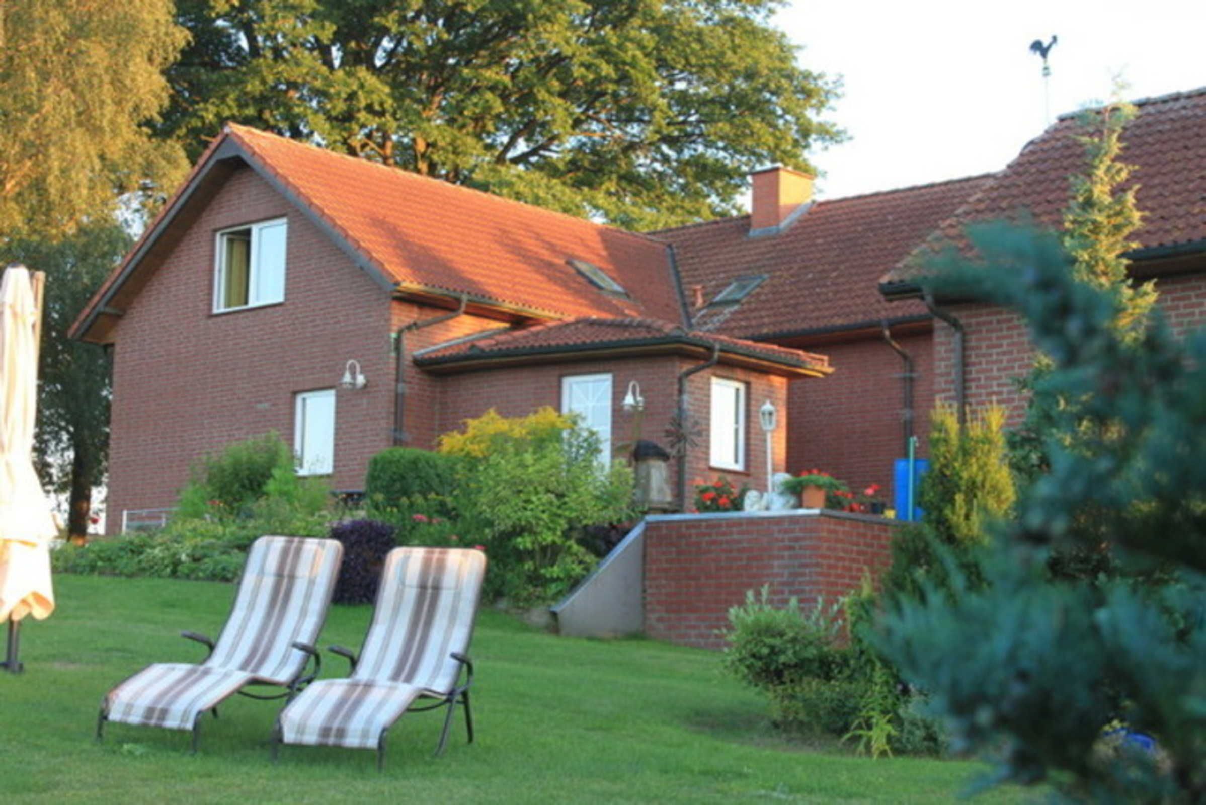 A two-story brick house with a red-tiled roof, a small garden with two striped lawn chairs, and a variety of plants and flowers.