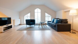 A spacious living room with a high ceiling, a flat-screen TV, a dark blue armchair, a grey sofa, a small coffee table, and a floor lamp. - Sea breeze - Villa on the beach (new build) (Heringsdorf)