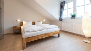 A bedroom with a wooden bed, a nightstand, a lamp, a window with curtains, and a radiator. - Sea breeze - Villa on the beach (new build) (Heringsdorf)