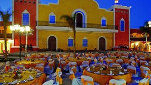 Une salle de banquet avec des tables dressées pour un événement formel, de grandes fenêtres et, en arrière-plan, un bâtiment imposant à la façade jaune et aux détails rouges.
