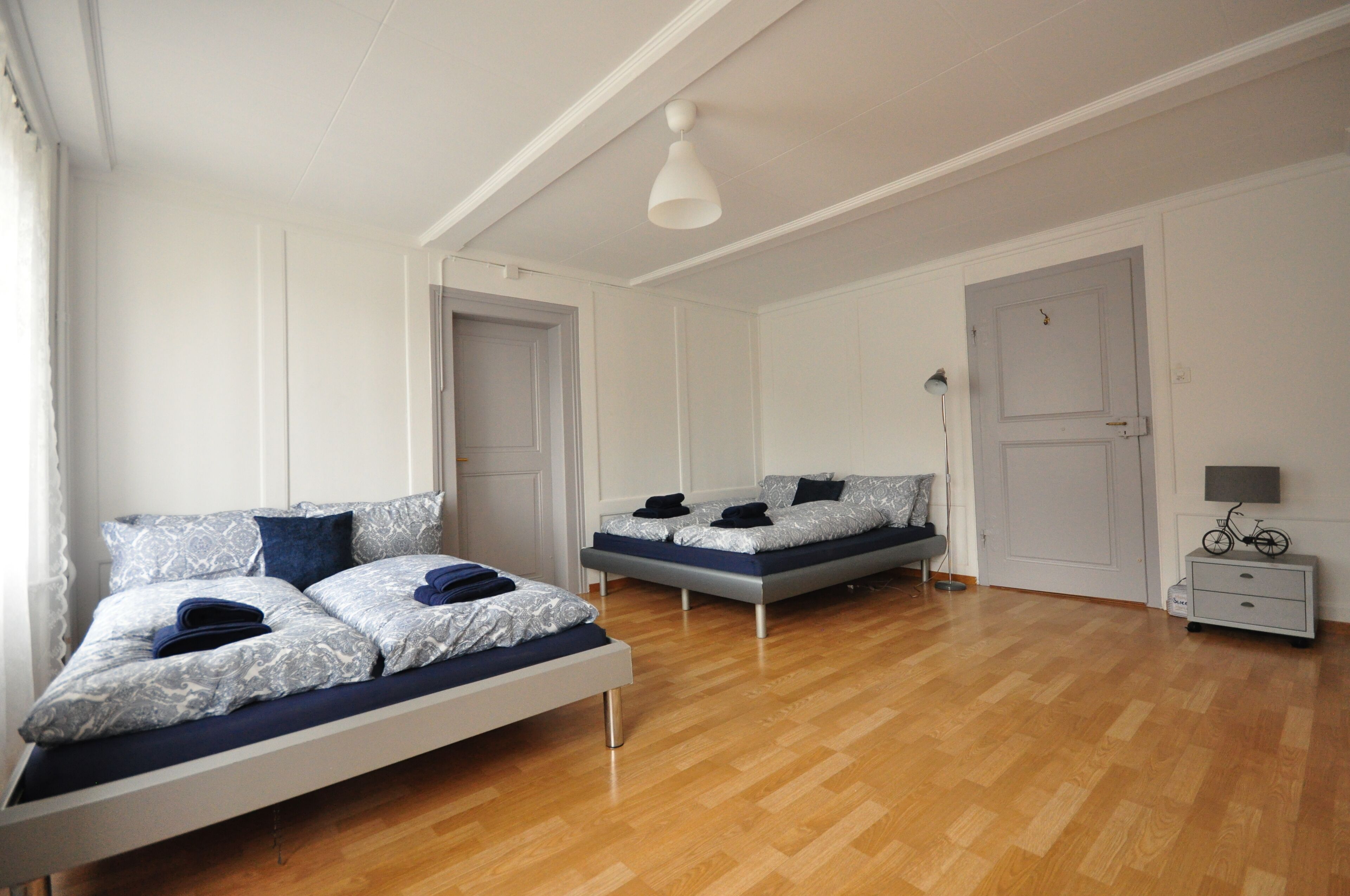 A room with two beds, a wooden floor, a white door, a small bedside table with a lamp, and a bicycle on the wall.