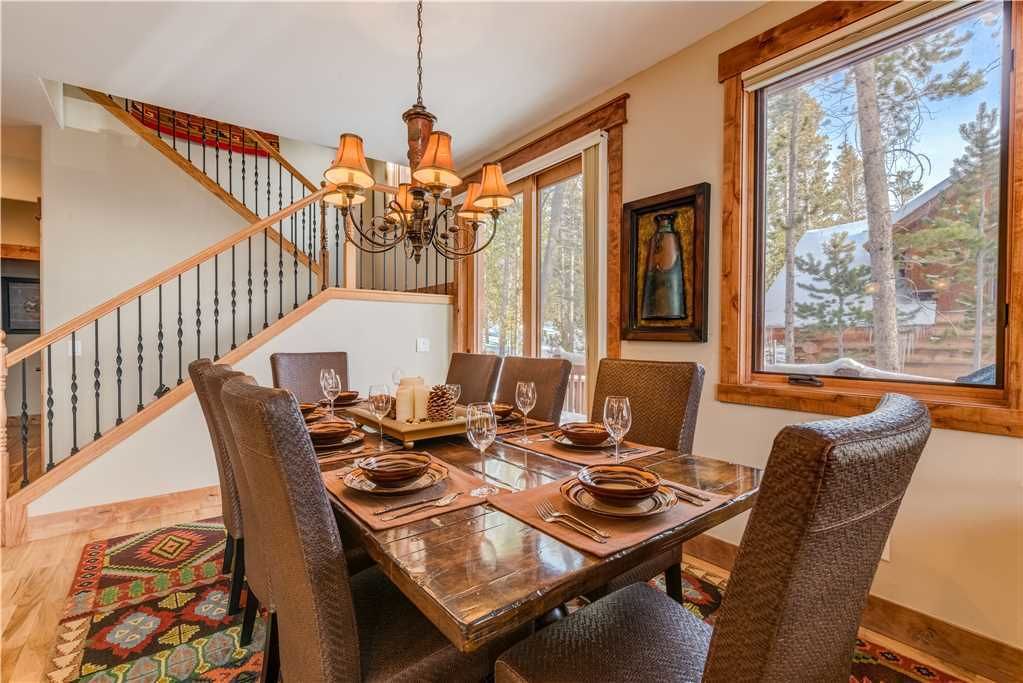 A dining area with a wooden table set for four, surrounded by chairs, a chandelier, and a staircase with a railing.