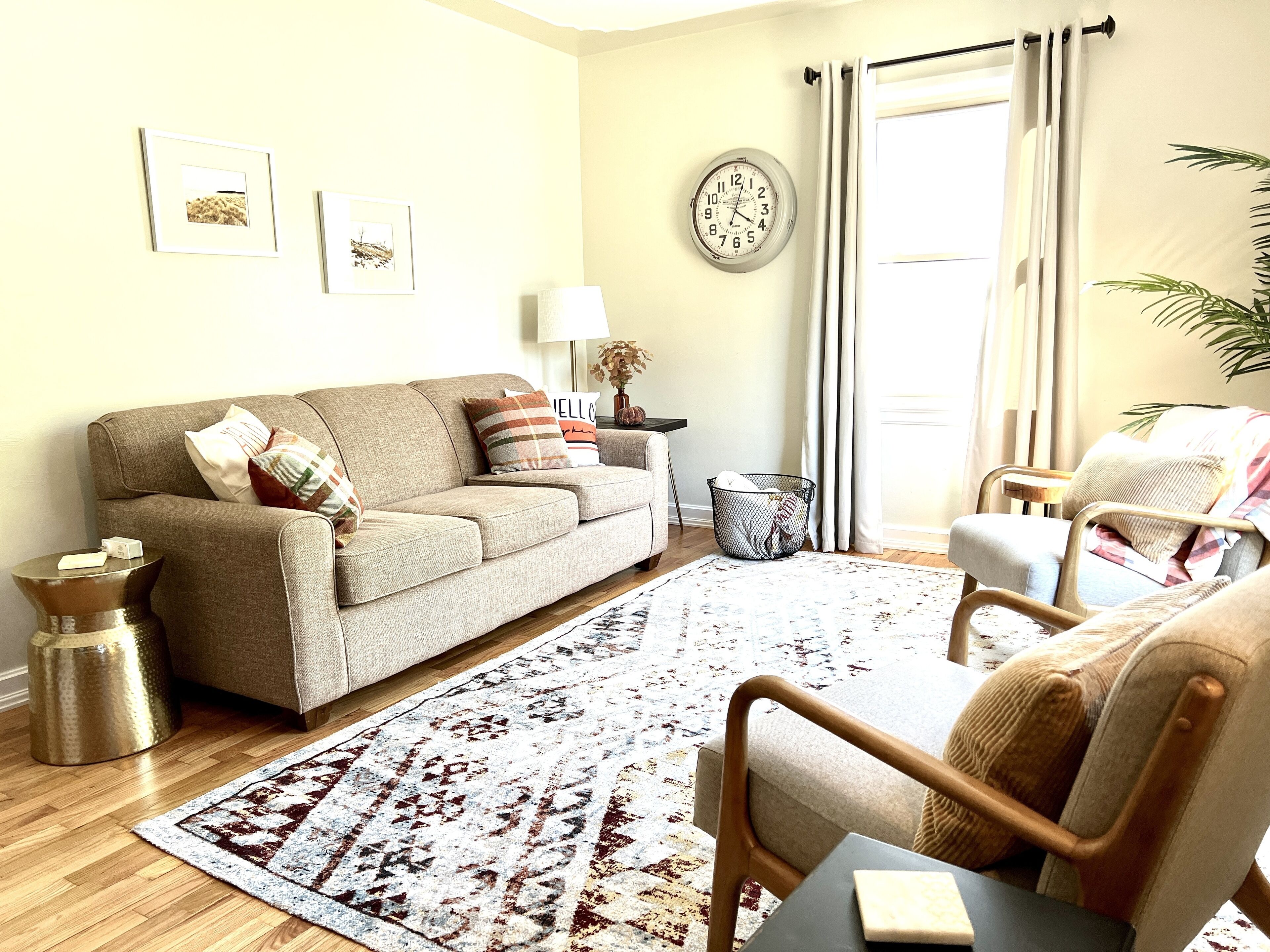 A living room with a sofa, armchairs, a patterned rug, a clock on the wall, and a window with curtains.