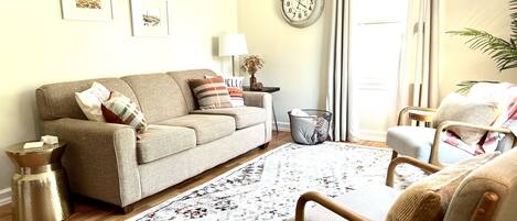 A living room with a sofa, armchairs, a patterned rug, a clock on the wall, and a window with curtains.