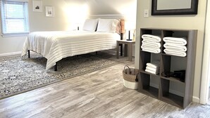 Une chambre à coucher comprenant un lit, une table de chevet, une commode, un miroir fixé au mur, une étagère en bois avec des serviettes, un tapis à motifs et une fenêtre avec des stores.