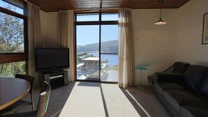 Ein Wohnzimmer mit Fernseher, einer Couch, einem Esstisch und Blick auf einen See durch große Fenster.