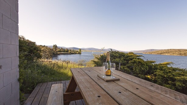Ein Holztisch mit einer Champagnerflasche und zwei Gläsern, mit Blick auf einen See und Berge im Hintergrund.