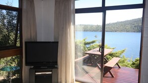 Ein Zimmer mit Fernseher, einer Schiebetür zu einer Terrasse mit Tisch und Stühlen sowie Blick auf einen See.