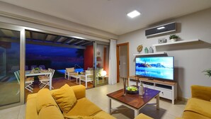 Ein modernes Wohnzimmer mit einem großen Flachbildfernseher, gelben Sofas, einem Holztisch und einer Schiebetür zu einem Essbereich im Freien.