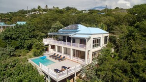 Una casa de dos pisos con piscina, rodeada de abundante vegetación.