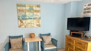 A room with two blue chairs, a small table, a television on a wooden cabinet, and a wall art piece with the words "Sunny," "Salty," and "Sandy." - Oceanfront condos with rare 2 night minimum rentals and linens included! (Ocean City)